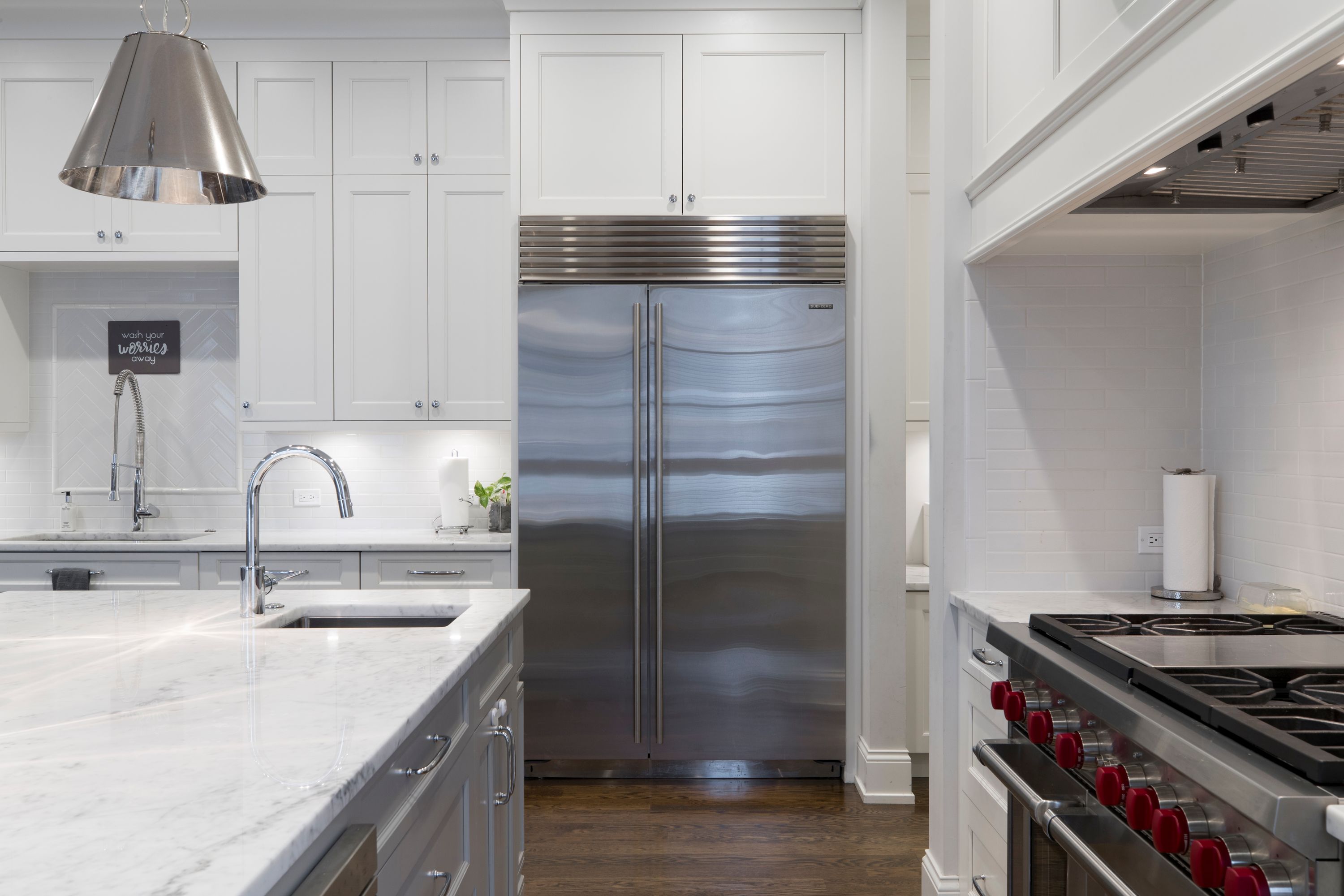 Tall built-in stainless steel refrigerator framed by custom white cabinetry, with marble counters, wood flooring and a professional range